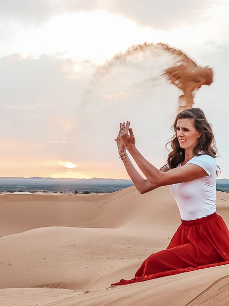 A woman in a red skirt tosses sand in Sahara Desert.
