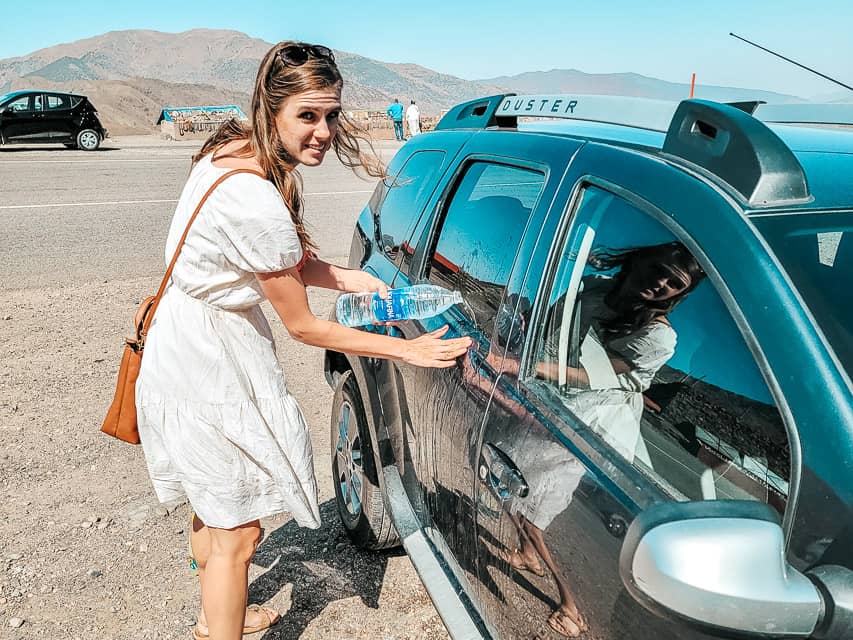 A woman tries to clean vomit off the side of a car with a water botte.