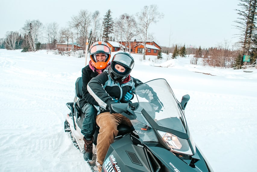Two people sit on a snowmobile with a cabin in the background.
