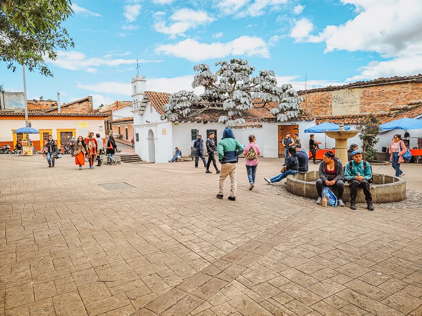 A small public square with cobble stones, a small fountain, and a little white church at the back. A few people are walking through the square.