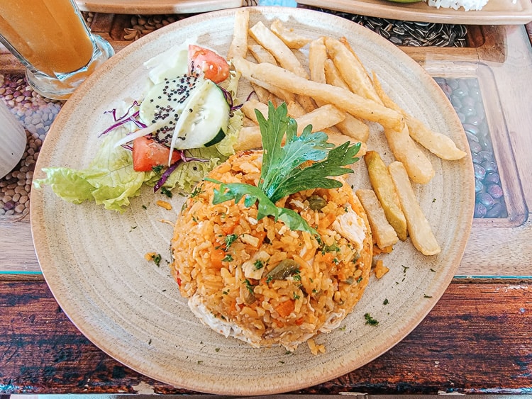 A plate of Arroz con Pollo. The rice is orange in color due to spices and mixed vegetables, combined with the chicken, and served in a mound. Side of fries and salad.