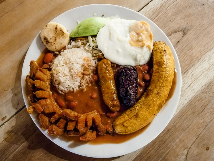 A plate of Bandeja Paisa, a traditional Colombian food consisting of sausages, rice, beans, fried egg, arepa, and plantain.