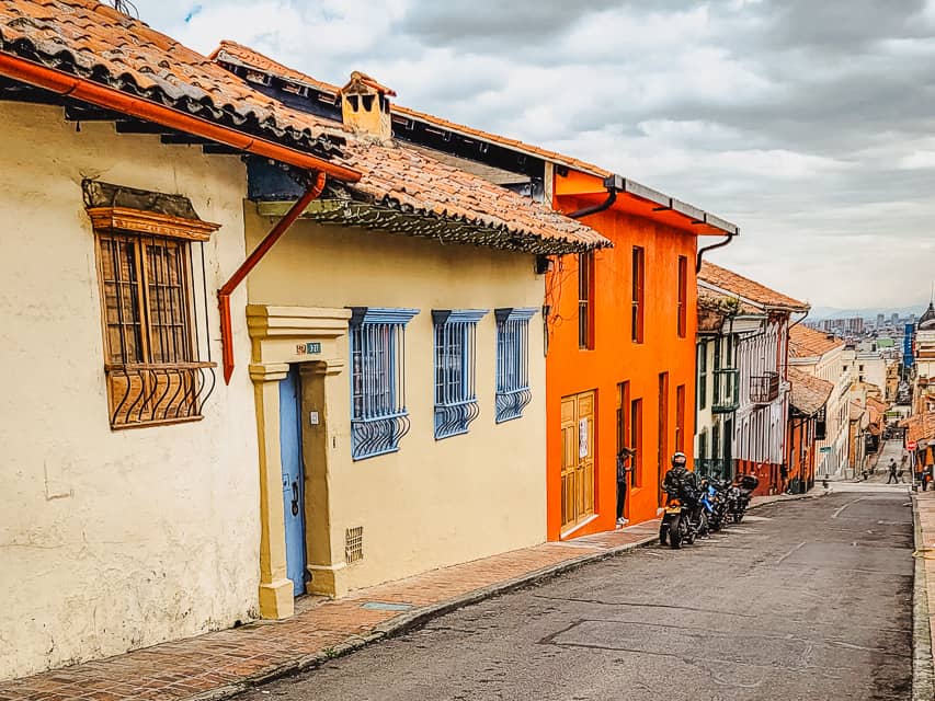 Colorful buildings, including beige walls, blue doors, orange doors, red walls, on a hilly street in Candelaria of Bogota - one of the best places to visit.