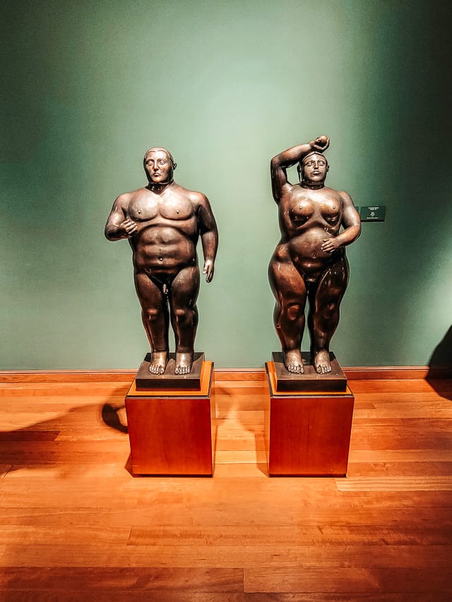 Two Botero sculptures are on display: a naked man and woman. The floor is hardwood and the wall is green.