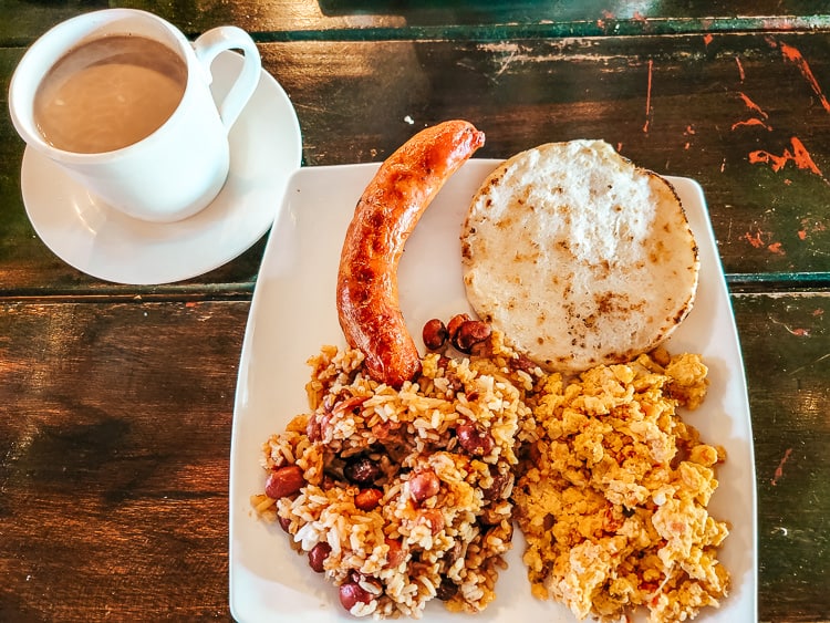 A traditional Colombian breakfast plate - arepa, scrambled eggs, rice and beans, and chorizo. A glass of hot chocolate is to the side.