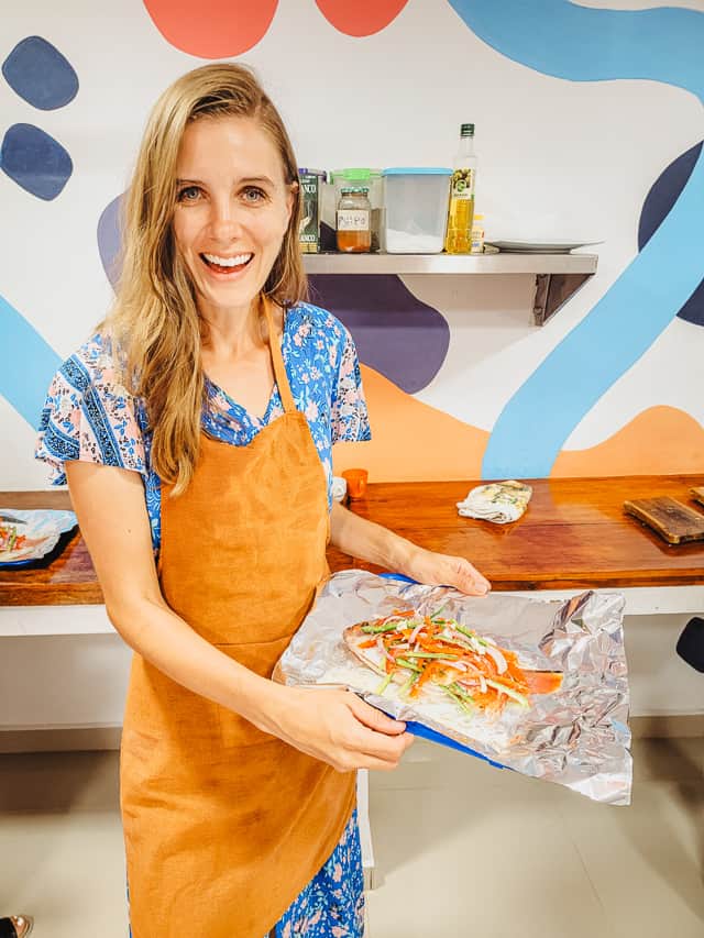 A smiling woman holds a tray with a raw fish rubbed with different spices and topped with vegetables. She stands in a kitchen in a cooking class in Cartagena.