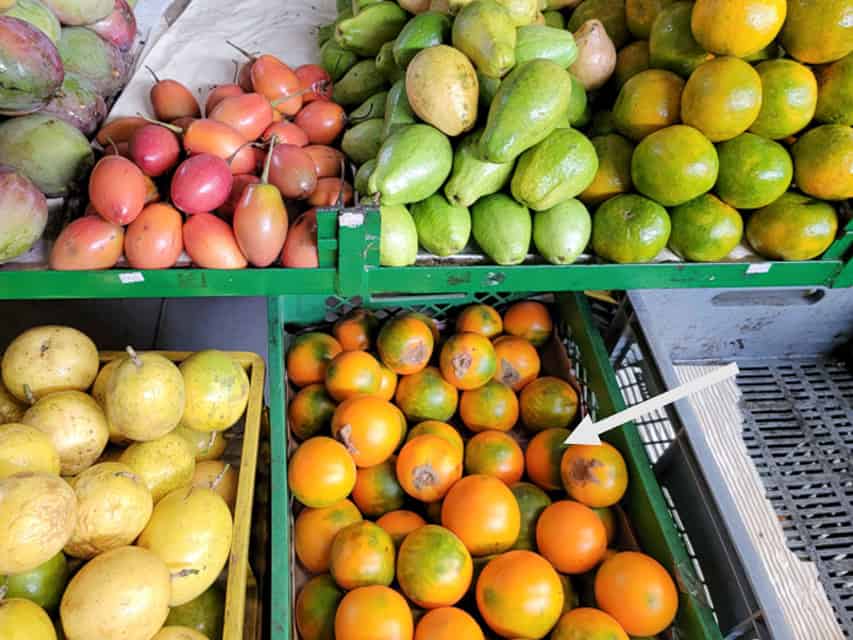 A selection of fruits at a Colombian market. A white arrow points to a crate of naranjilla. Naranjilla (lulo) has a thin, rough, orange rind. It tastes similar to kiwi.