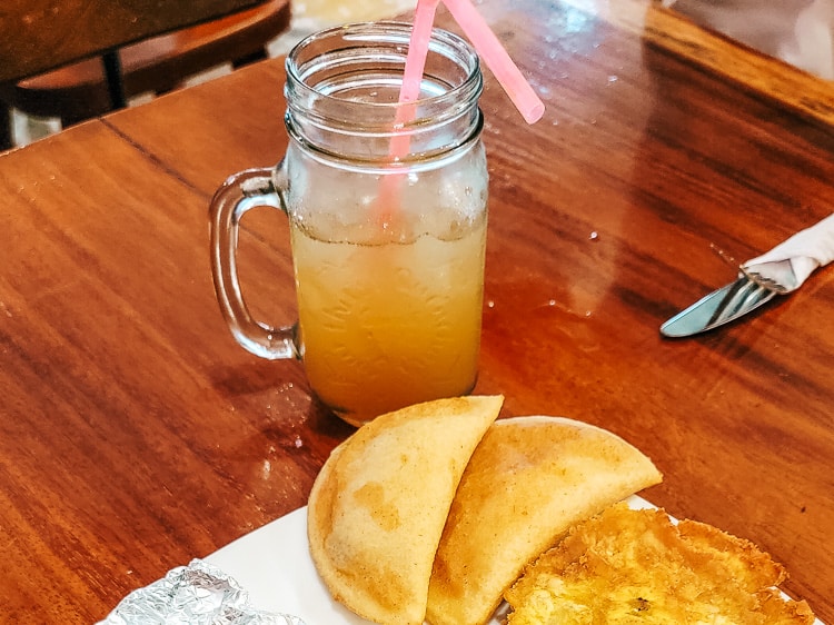 A mason jar full of aguapanela, a tan drink based on raw sugar cane. Empanadas and patacones are served nearby.