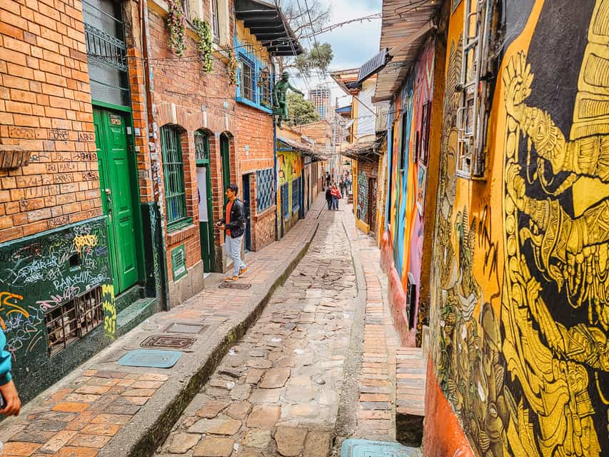 A narrow alley, with cobblestoned street, brick buildings, and some colorful murals and street art on the walls. A few people walk through.