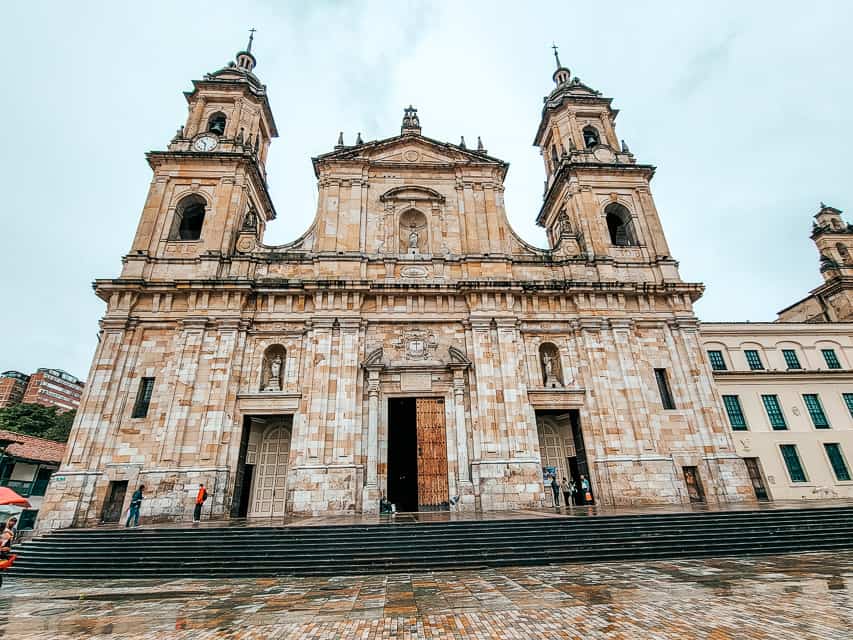 The exterior of the Primada Cathedral of Bogota, standing on a ledge with 10 steps up, two spires, 3 entrance doors on the front. It's made of beige stone.