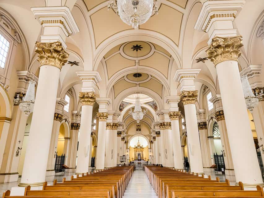 The inside of the Primadial Cathedral of Bogota - it's very white and tan, with columns, arches, intricate chandeliers, and wooden benches.