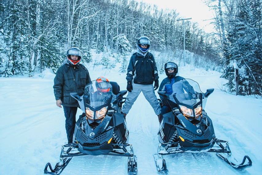 Four people with two snowmobiles in the woods.