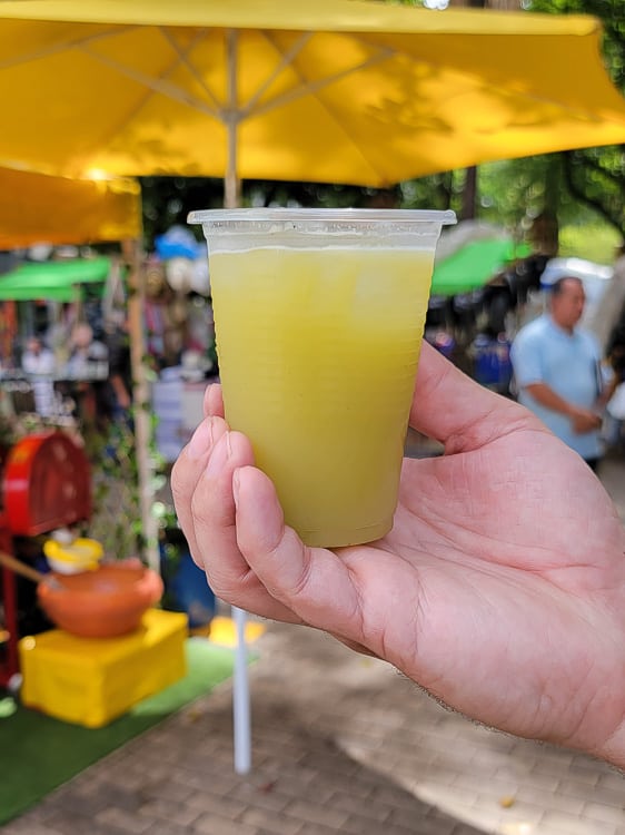 A glass of bright yellow sugar cane and lime juice in a plastic cup. There is a market in the background.