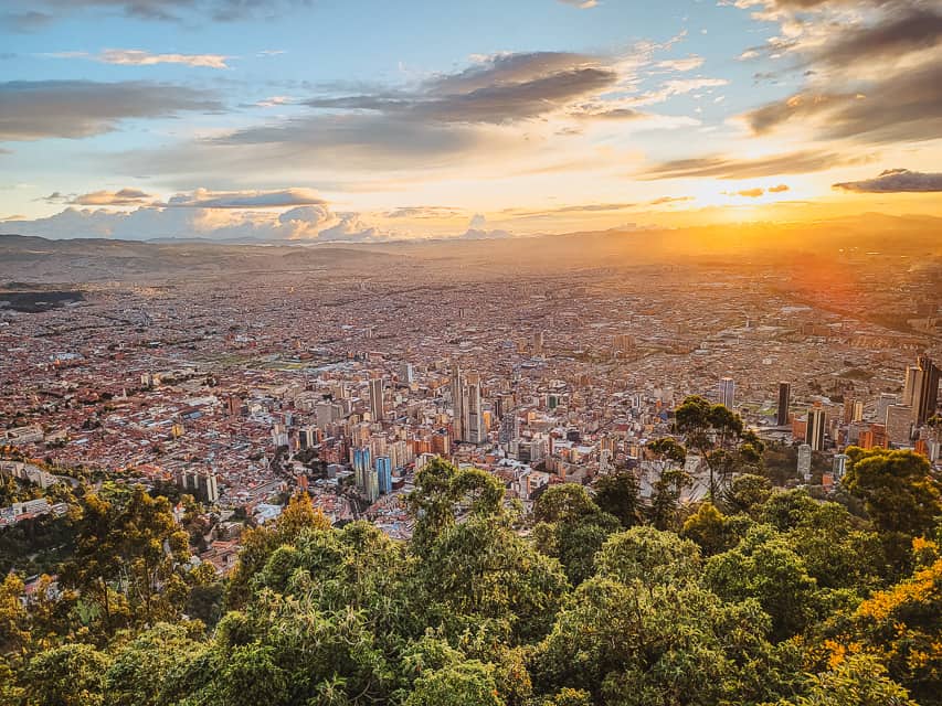 The sunset lights up the city of Bogota, with skyscrapers and other buildings present. Greenery from the mountain is in the foreground. Admiring this view is one of the best things to do with 3 days in Bogota.