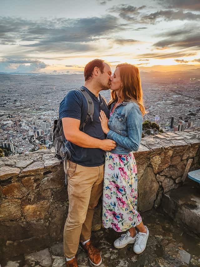 A man and a woman kiss in front of a ledge on a mountain overlooking the city of Bogota. The sun sets in the background.