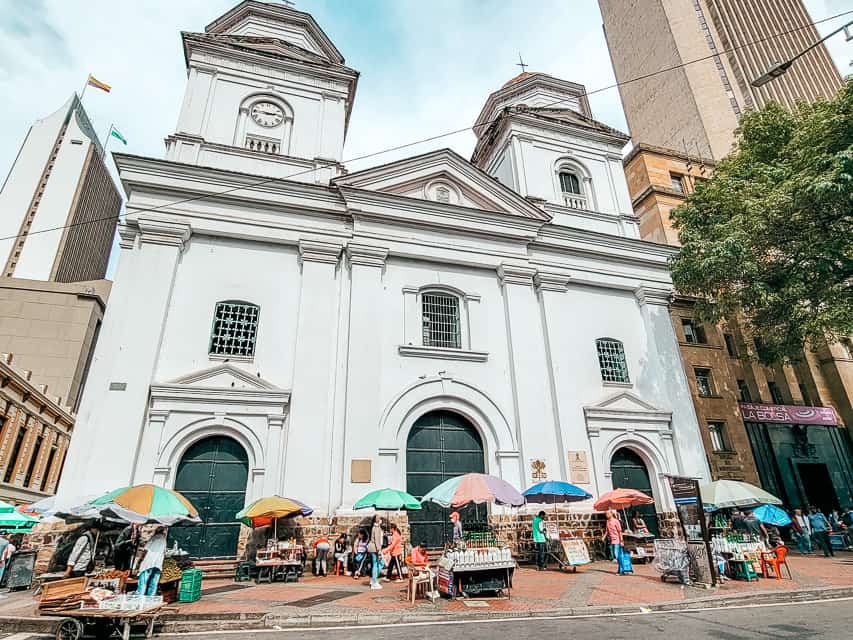 A white stone colonial church standsin the middle of the sidewalks with many vendors outside the doors selling food.