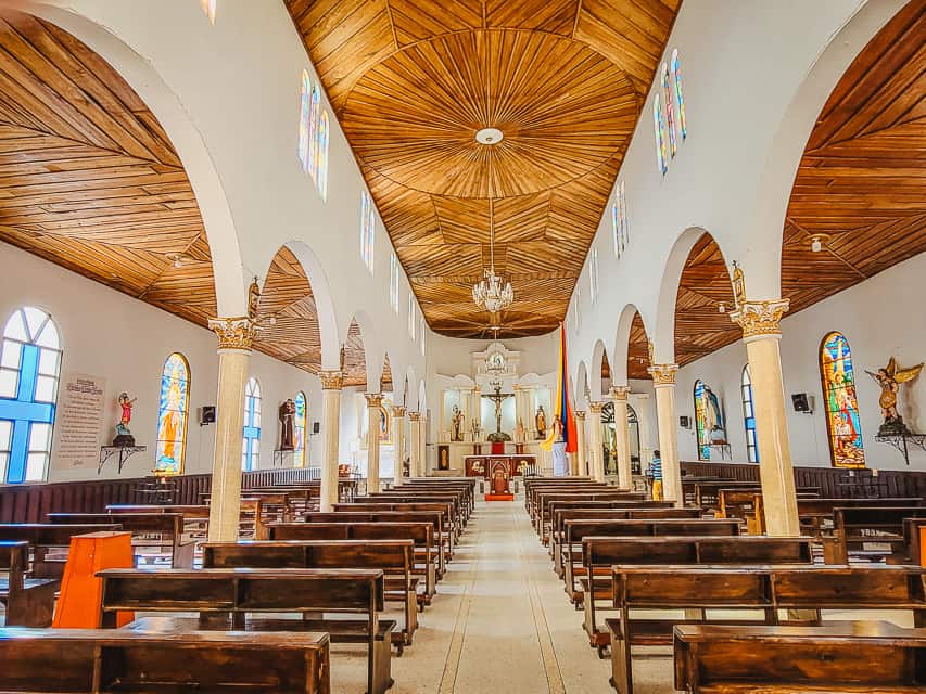 The inside of the Our Lady of Carmen church in Salento, with tiled floors, probably 15-20 rows of dark wooden benches, stained glass windows on the exterior walls, a ceiling of planked wood in different designs, and archways leading up to the front of the church. This is one of the most beautiful places to visit in Salento.