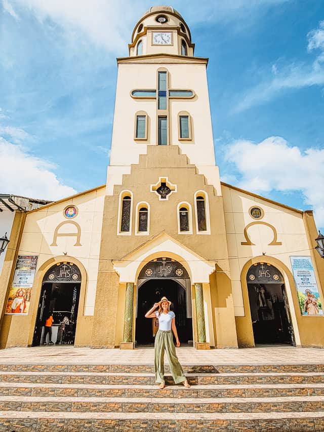 A woman in a straw hat, white shirt, and green, flowy pants stands on some steps leading up to the church in Salento. The church is tan and beige, has three entrance doors, a tall steeple, and a cross on the tower.