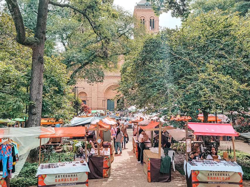 A market with lots of huts with colorful tops. There are an assorment of displays and lots of people shopping around. It is located in the middle of a park with trees everywhere.