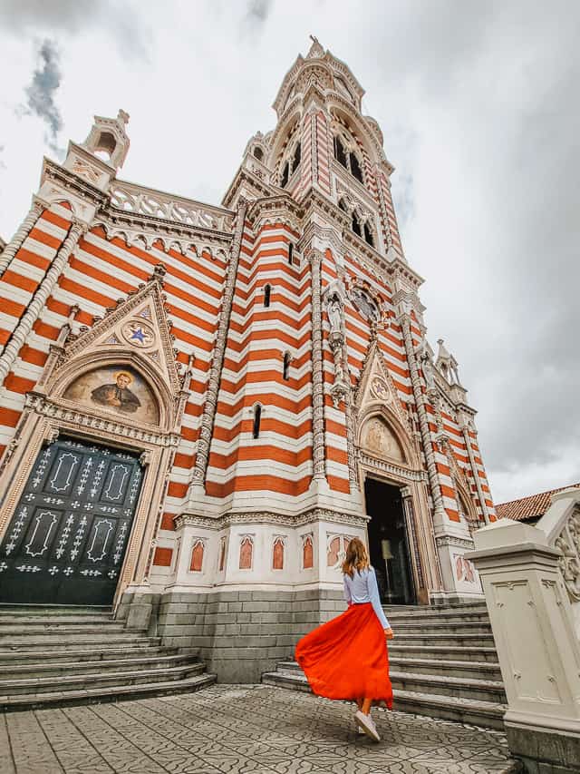 A woman in a white shirt and red skirt walks up to a church constructed in rows of alternating red and white bricks (the candy cane church).