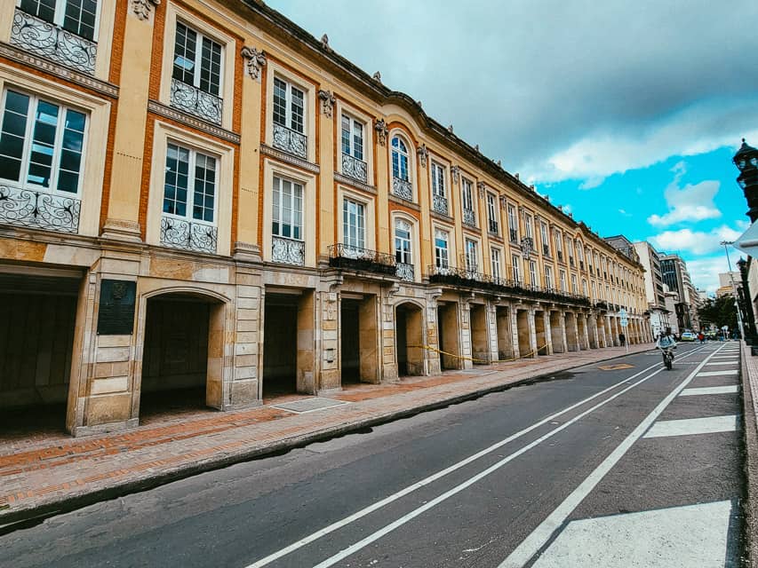 A fancy street in Bogota, with an archway on the street level, a golden building, with windows trimmed in red bricks, and a fancy cast iron balcony.
