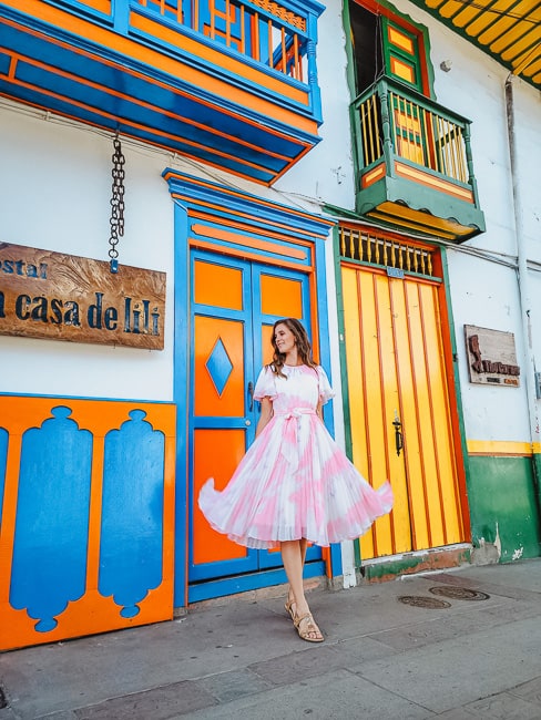 A woman in a pink and white dress stands smiling in front of a door with orange and blue, with a door in yellow, green, and orange next to her.