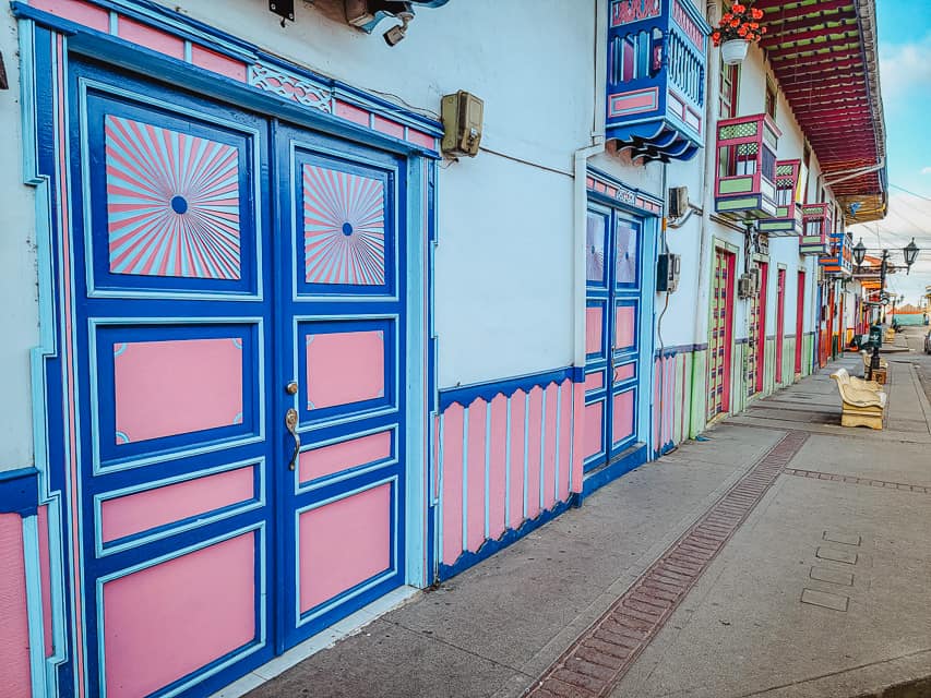 This is a picture of calle real. You can see one side of the street, with several different shops (and the subsequent different color schemes) in them. Closest to the viewer are doors and chair railing with pink, light blue, and dark blue coloring, then there's a shop with light and dark green and red coloring, and farther you can see doors and windows with red, blue and orange. Calle Real is one of the most beautiful places to visit in Salento, Colombia.