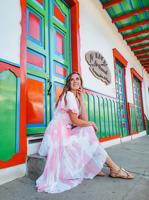 A woman in a pink and white dress sits on a step in front of some shops. The doors, chair railing, and roof overhang are painted in bright blue, red, and green colors.
