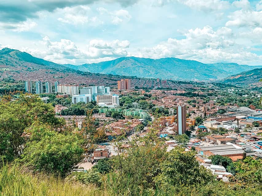 The view from the top of a mountain with lots of plants throughout the beautiful city in the valley of lots of mountains.