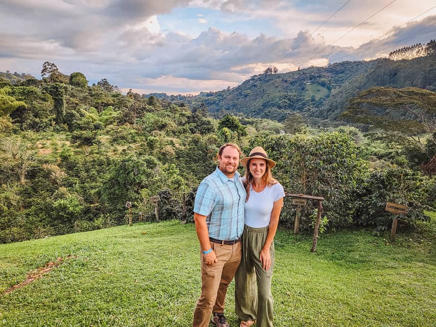 A man in a blue shirt and tan pants, and a woman in a white skirt, green pants, and straw hat smile at the camera on a small hill in front of a lush valley in the background.