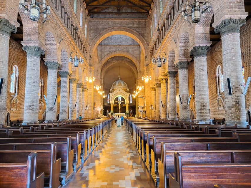 A room with lots of wooden pews leading up the room with white stone pillars leading that way. There are lights hanging from the ceilings also leading up towards the front