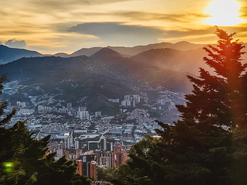 The view from a mountain at sunrise with the sun reflecting on the buildings and pine trees. There are continuing mountain ranges going behind the city.