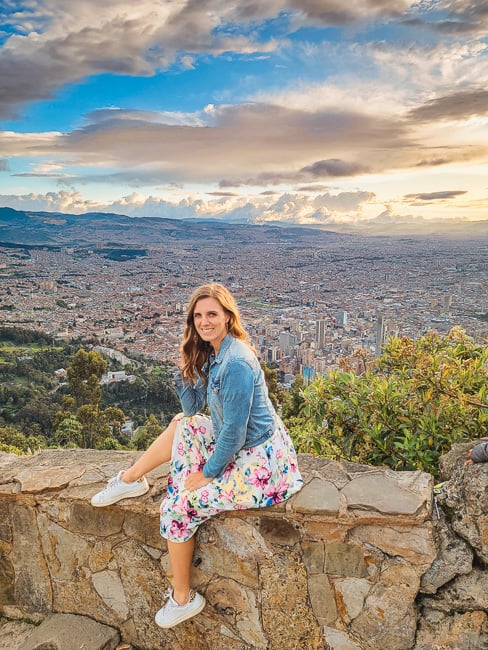 A woman in a flower dress and jean jacket sits on a ledge on a mountain that overlooks a valley and the city of Bogota.
