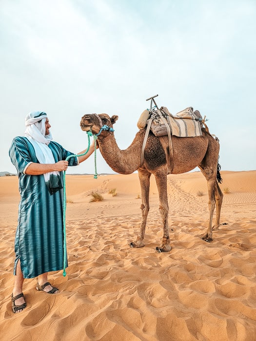 A man in a blue and black striped kaftan and a white turban looks at a a camel in the Sahara desert, a unique and incredible thing to do near Marrakech.