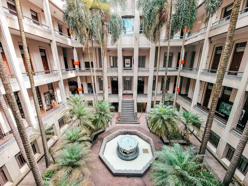 A hotel with many rooms on 3 different stories, on the main level there are palm trees and bushes surrounding a blue water fountain