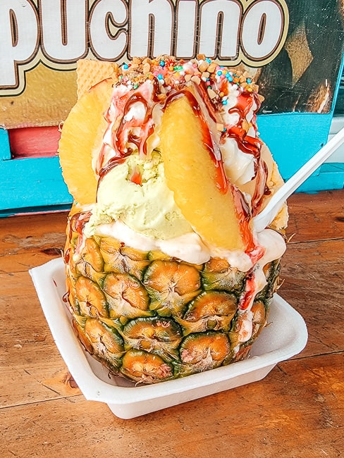 A pineapple is stuffed full of ice cream, fruit, and drizzled with a syrup and sprinkled with sprinkles.