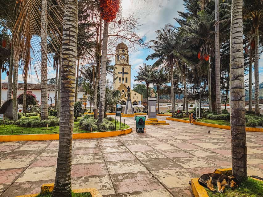 The town square, with large square concrete brisk, several raised areas with grass and palm trees, a small statue in the middle, and a church behind it. Visiting the square is one of the best things to do in Salento.