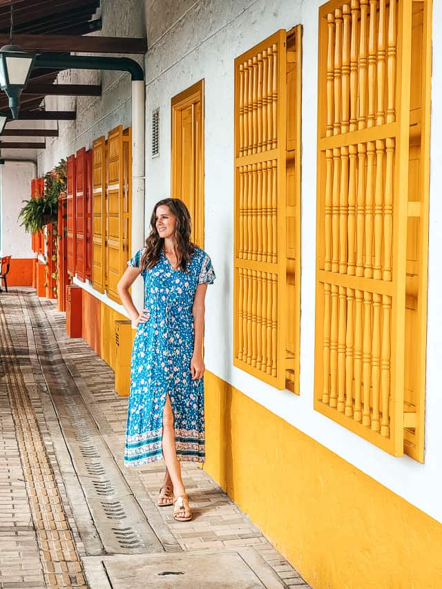 A woman in a blue floral dress walks past a white and yellow house with yellow shades.