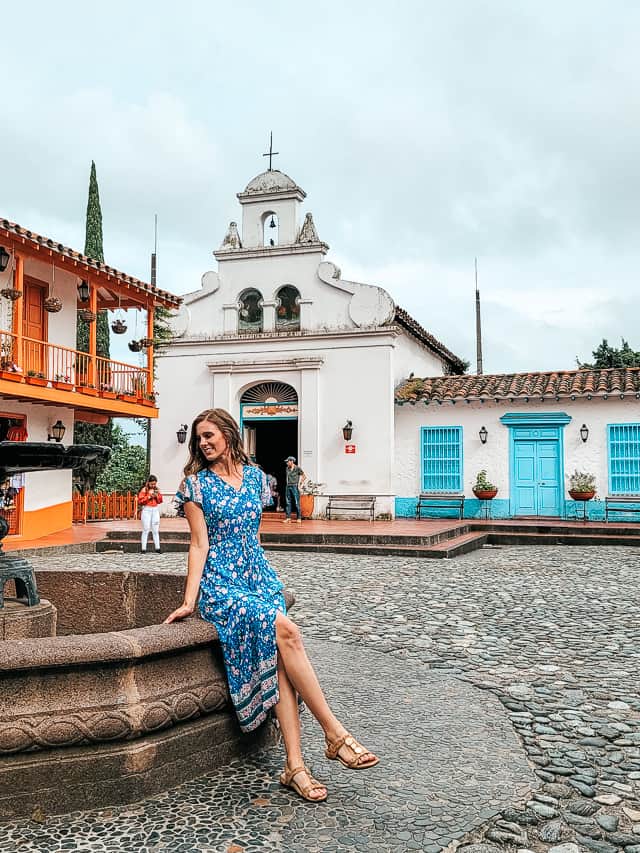 A woman sits on the edge of a stone fountain in the middle of a stone square with colorful buildings surrounding the perimeter.