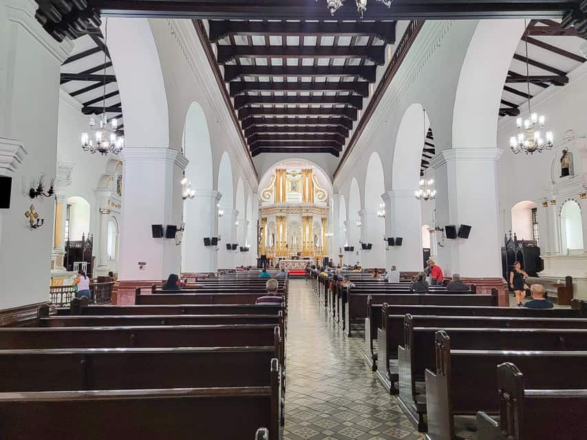 A very pristine church with white pillars and dark brown wood pews leading up to the gold altar.