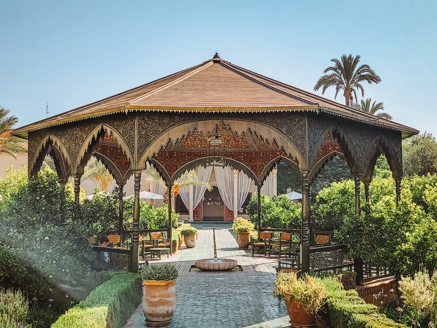 A beautiful, large dark wood gazebo with a decorated interior roof stands in the middle of a garden, with edges and plants surrounding it, tiled walkways going through it, and columns in the background.