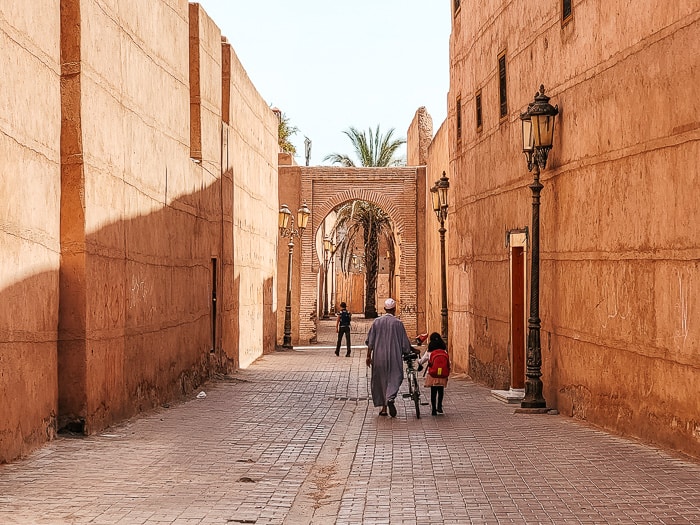 A view down a lesser known spot in the medina - with a Moroccan man in a kaftan walking a hike, red building walls, and an archway in the distance.
