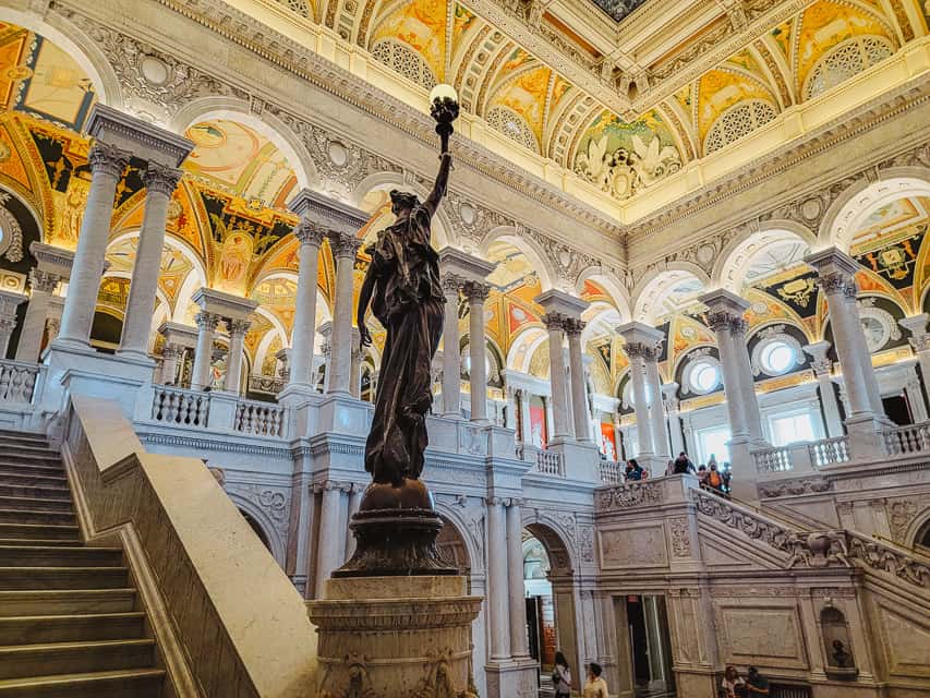 A bronze statue of a woman holding a torch stands in the main atrium of the Library of Congress with columned porticos lining the room and an intricately designed ceiling.