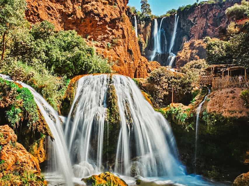 A view of Ouzoud Falls - water streams down multiple ledgs of dark, vibrant red rock, with some bushes and trees lining the riverbanks. This is one of the most unique things to do from Marrakech.