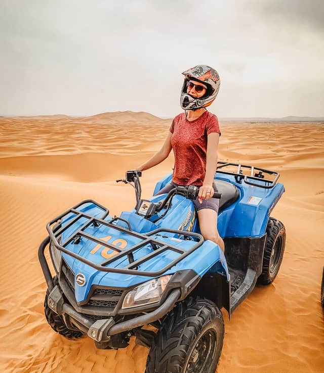 A woman wearing a helmet and sunglasses sits on top of an ATV, the rippling dunes of the Sahara stretching out behind her.