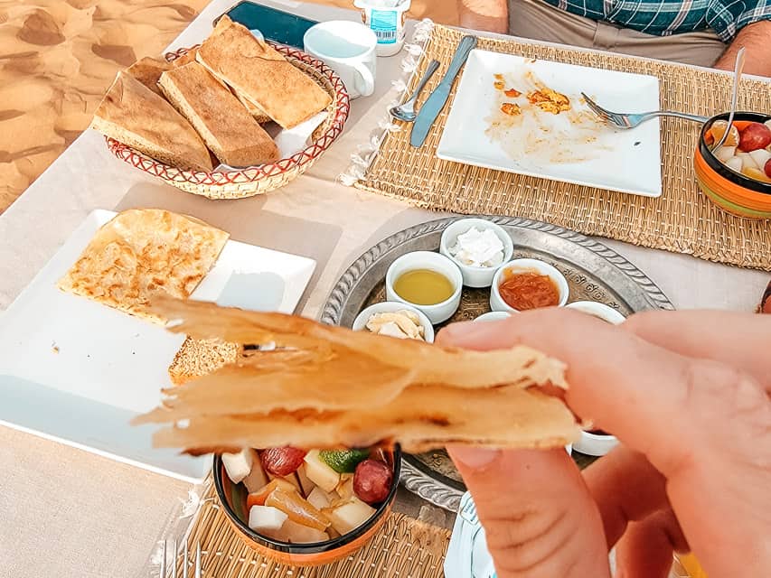 A hand holds a section of Moroccan crepe close to the camera with other crepes, breads, and various toppings on the table. A wonderful Moroccan breakfast food.