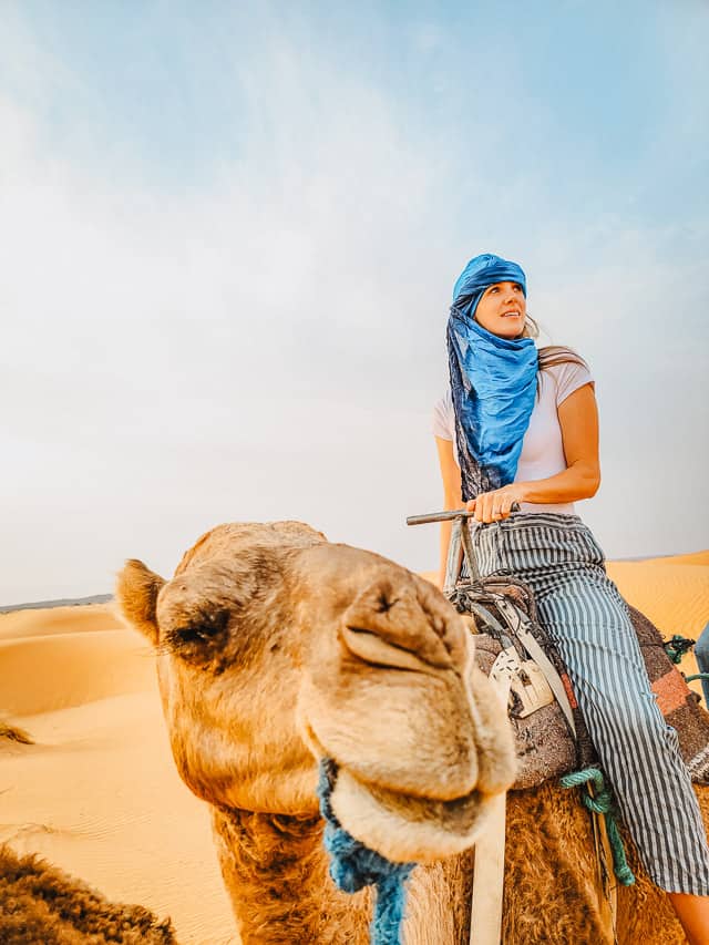 A woman riding on the back of a camel with the camel's face pointing towards the camera in the middle of the Sahara sand dunes.