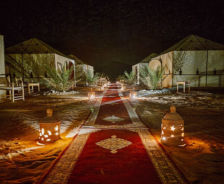 A typical Sahara desert tour luxury camp at night - a line of long, skinny, red carpets make a pathway down the center of the camp, with lanterns placed alongside the carpets. Tan, square shaped tents with a pointed top line linel the carpet, and greenery is planted at intervals.