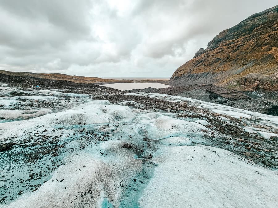 The view of the landscape around the blue ice glacier with a grass and rock hill with a small lake in the distance.