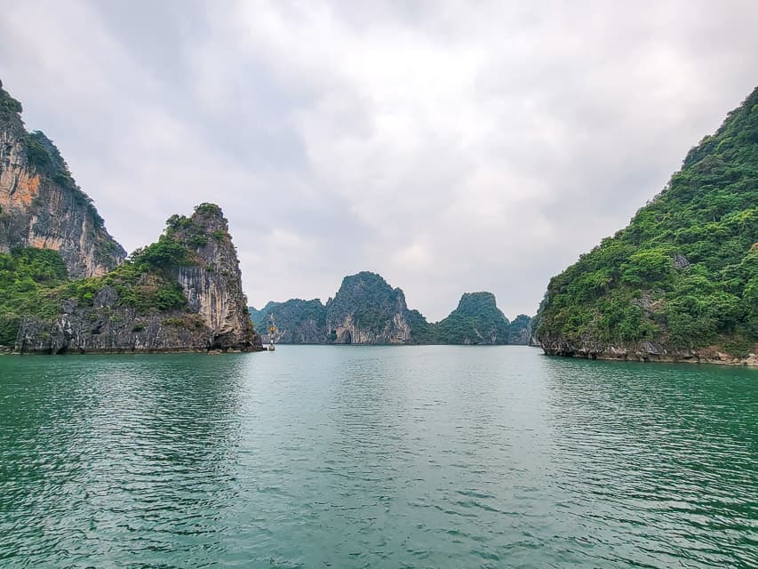 The greenish blue water of Halong Bay is in the foreground, with several ridges of mountain karsts, some covered with greenery, are in the background.